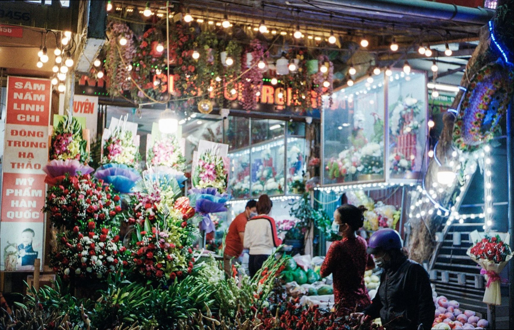 Animado mercado de flores nocturno con coloridos arreglos florales y compradores.